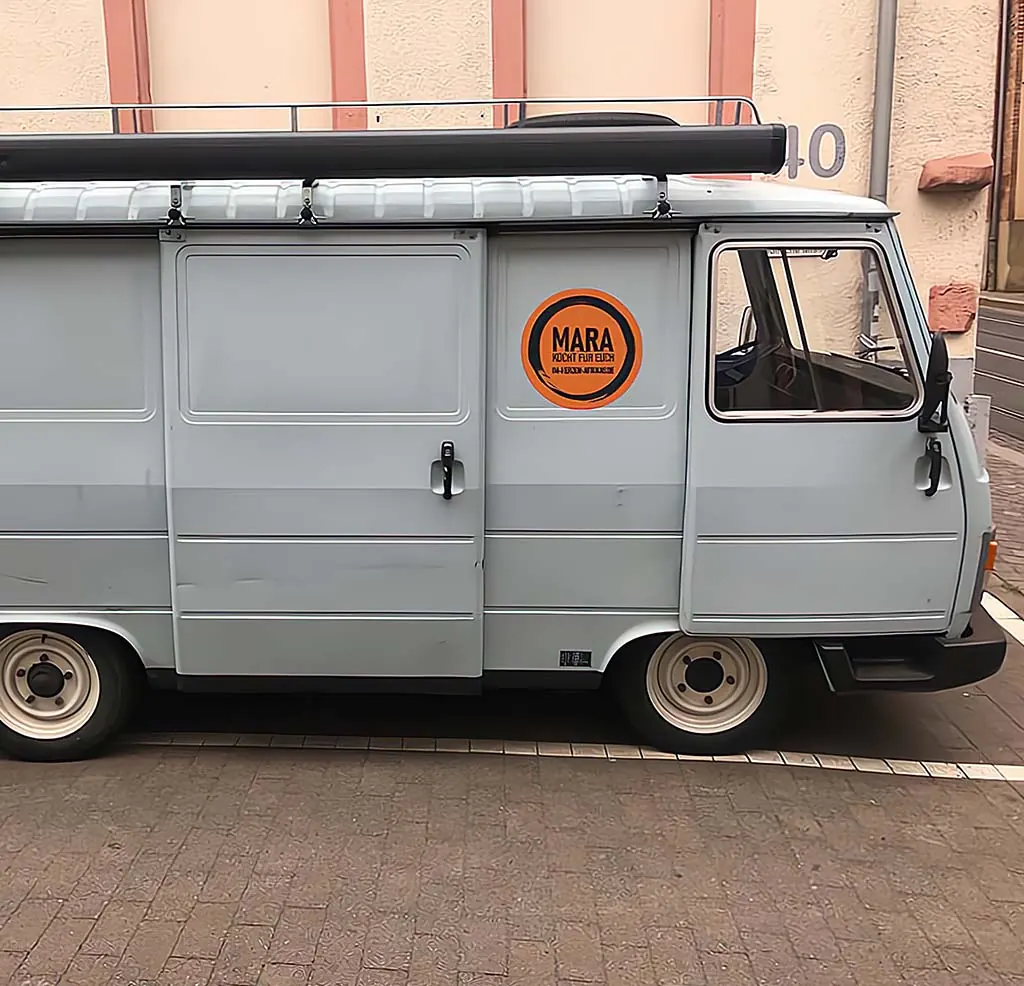 Hellgrauer Kleinbus mit dem orangen Zeichen „Mara kocht für euch“