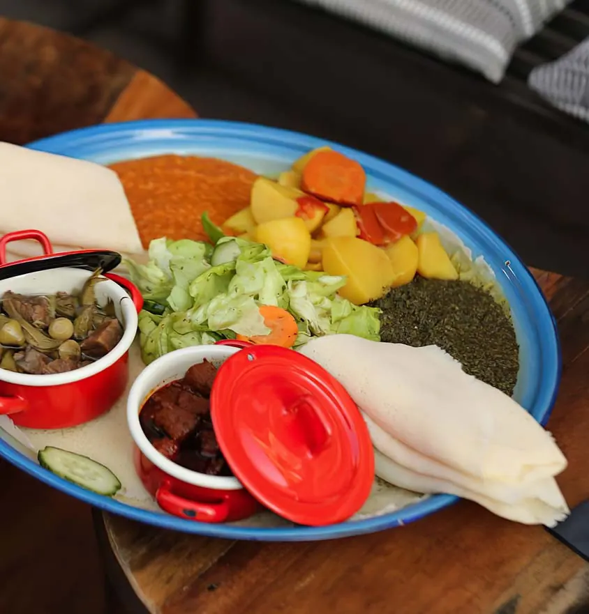 Blaue Emaille-Platte mit Injera, Fleisch in roten Töpfchen, Salat und Gemüse