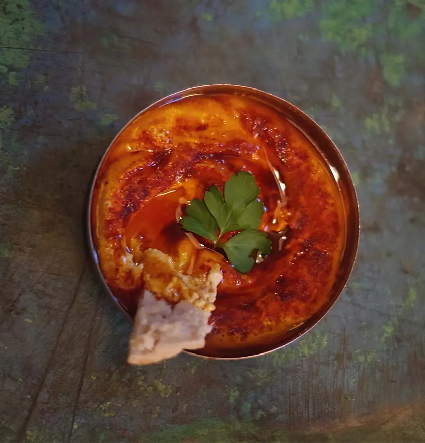Hummus in einer Metallschale mit Olivenöl, Berbere und Petersilie, daneben ein Stück Fladenbrot